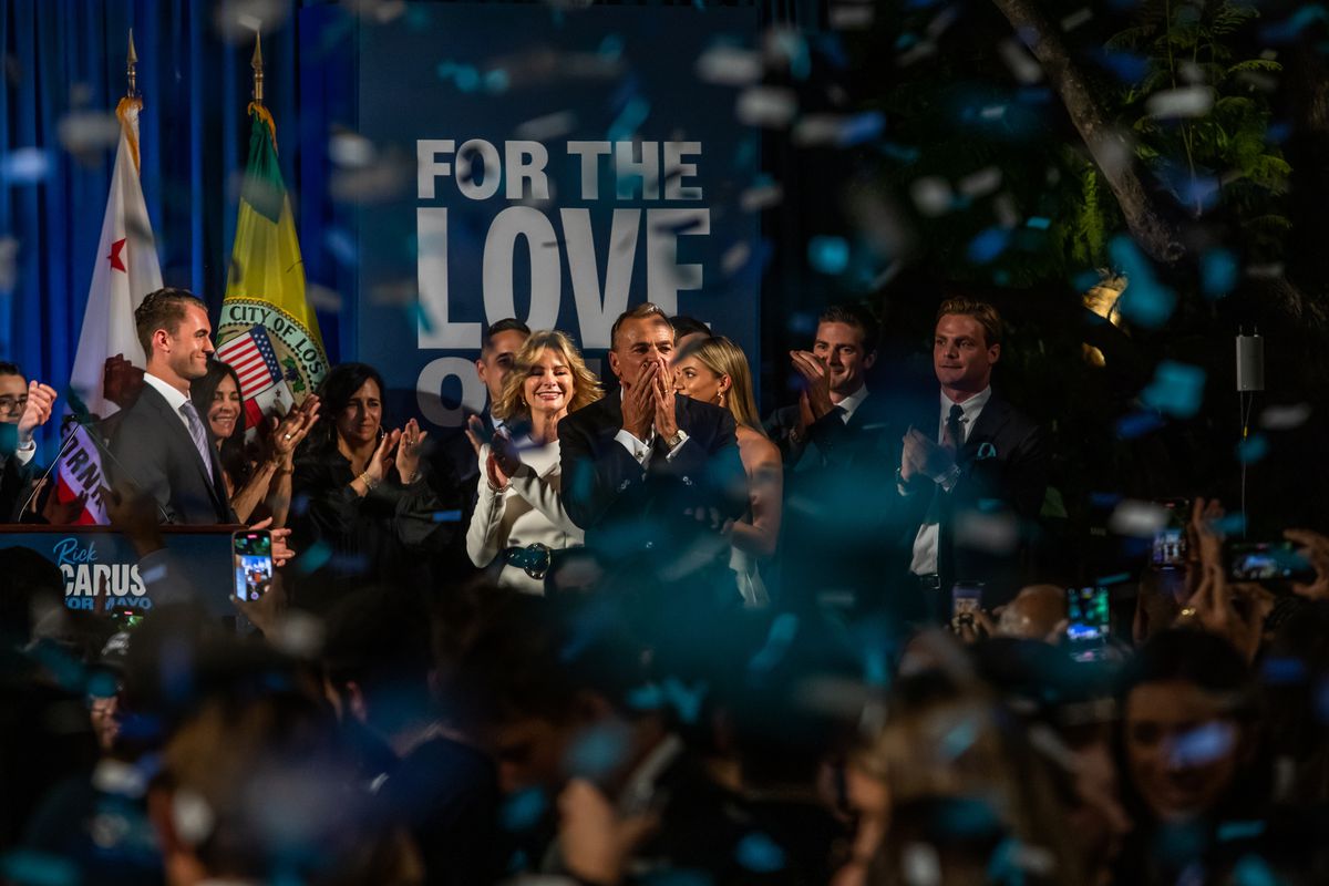 Caruso, in a dark suit and surrounded by family and supporters on a stage, covers his mouth as if about to blow a kiss to the cheering crowd that populates the foreground of the photo. Blue confetti rains from the ceiling. A sign behind Caruso reads, “For the Love of LA.”