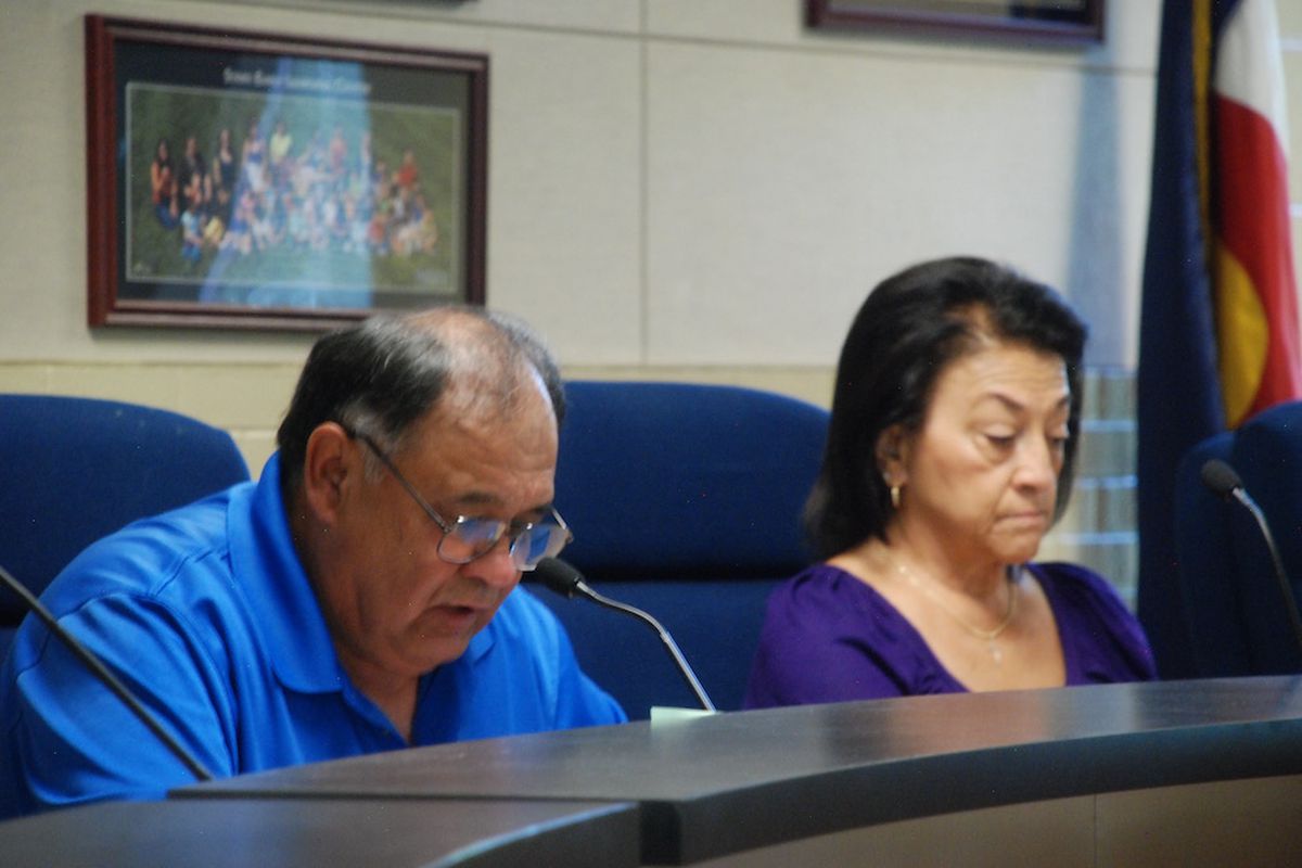 A man wearing glasses in a blue shirt and a woman also wearing a blue shirt sit at a table. Both are looking down. 