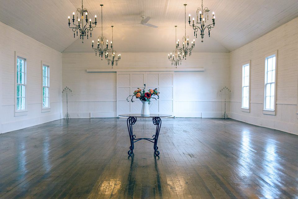 Big event room with whitewashed walls, chandeliers, and a small table with floral arrangement in the middle