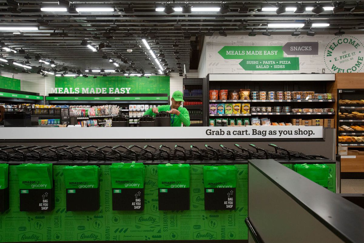Green grocery bags and shopping carts in the foreground; in the background, a Amazon Go Grocery worker, along with shelves of goods.