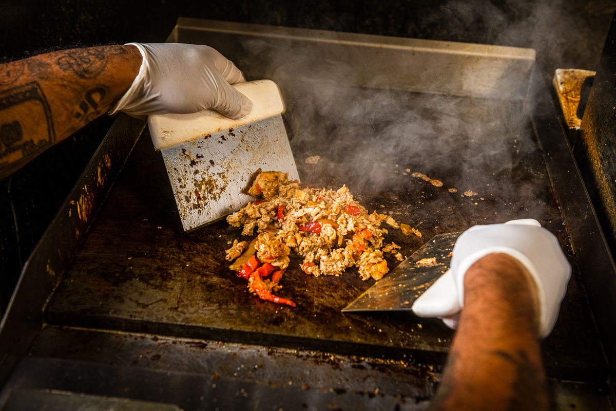 A pair of gloved hands chop the chicken and peppers on a flat-top grill.