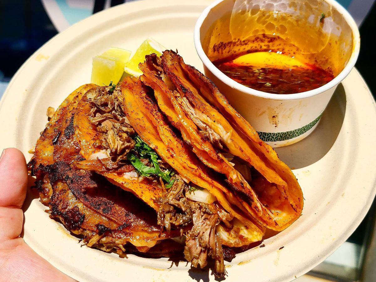 Three crispy tortillas filled with juicy meat, stacked on a paper plate beside a disposable cup of consomme