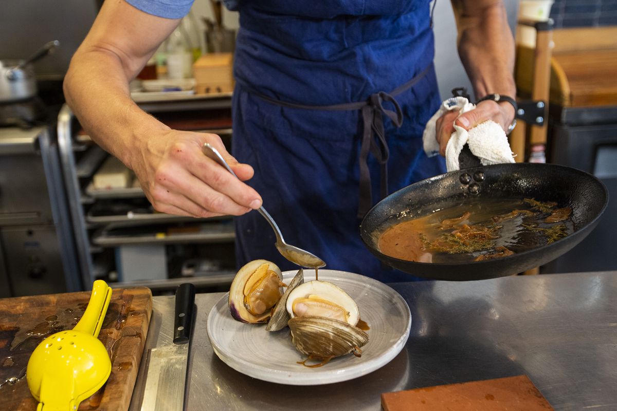 A man in a blue apron spoons sauce over a clam dish on a white plate.