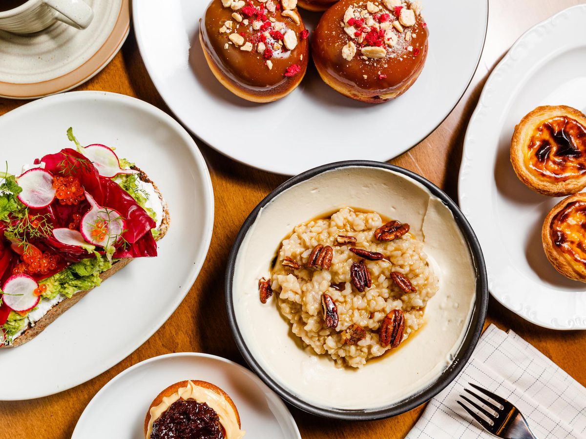 Atop a wooden table, a spread of five plates, including doughnuts, egg tarts, toast, and brown rice porridge.