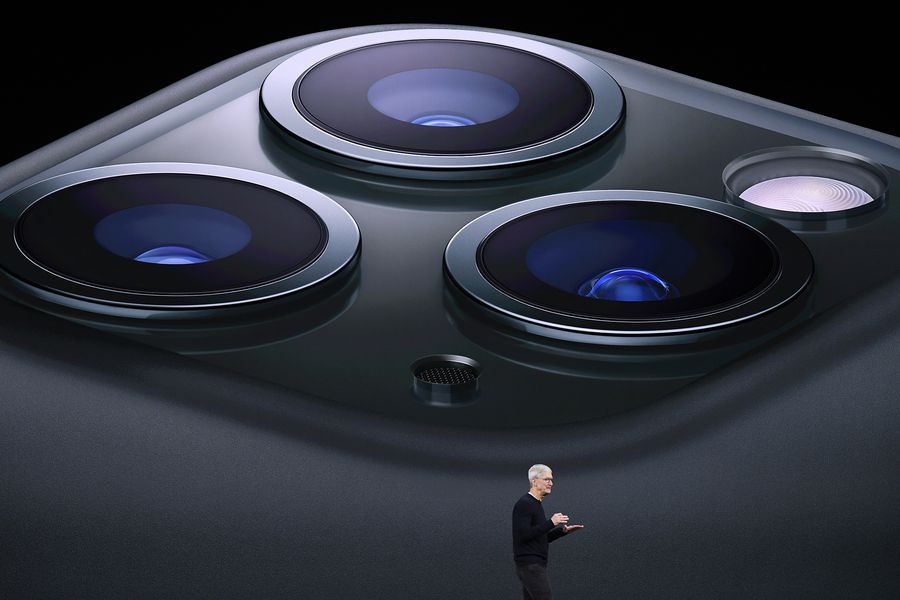 Tim Cook onstage at an Apple event with a huge picture of the three camera lenses on the back of an iPhone projected behind him.