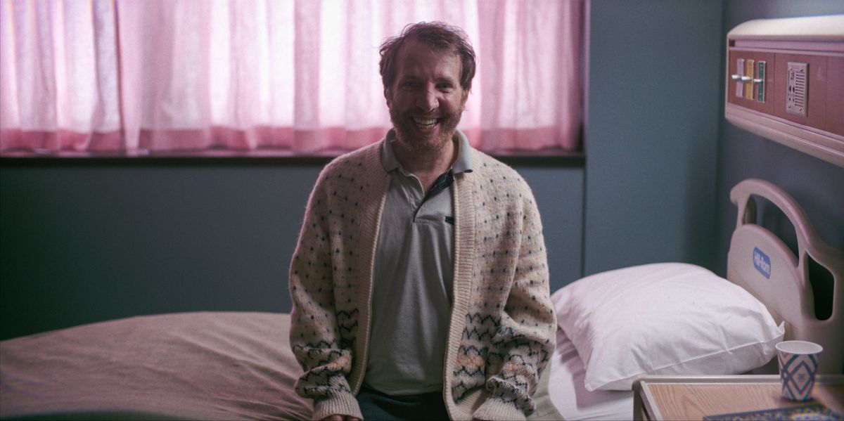 A red-haired bearded man in a sweater sits on a hospital bed in front of pink curtains with the biggest smile ever