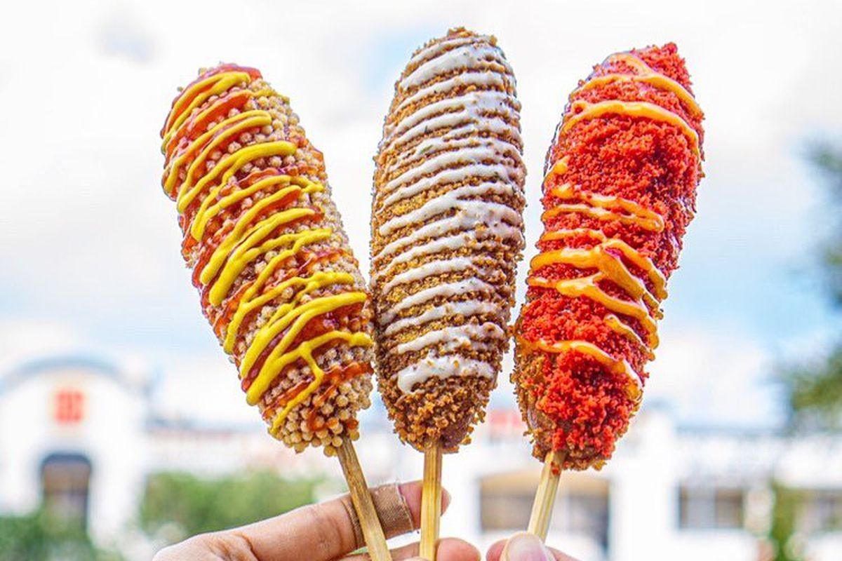 a pair of hands holding three elaborately-decorated corn dogs with different toppings