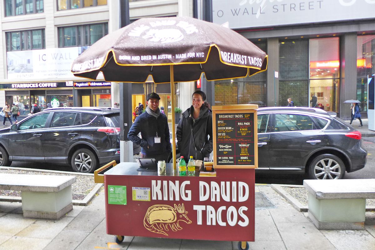 Two people stand under an umbrella at a small red food cart, whose sign reads “King David Tacos” in capital letters