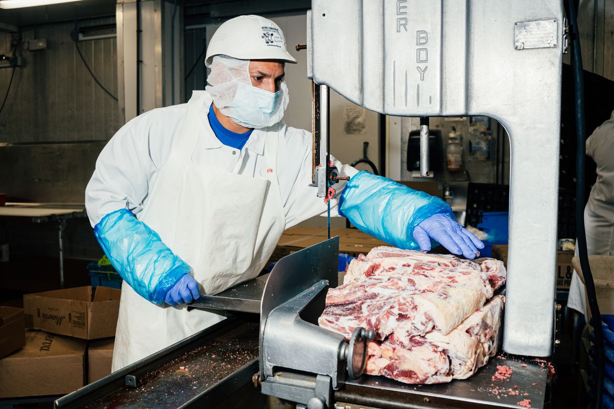 A man dressed in whites, a mask, and gloves working with meat at a meat processor.