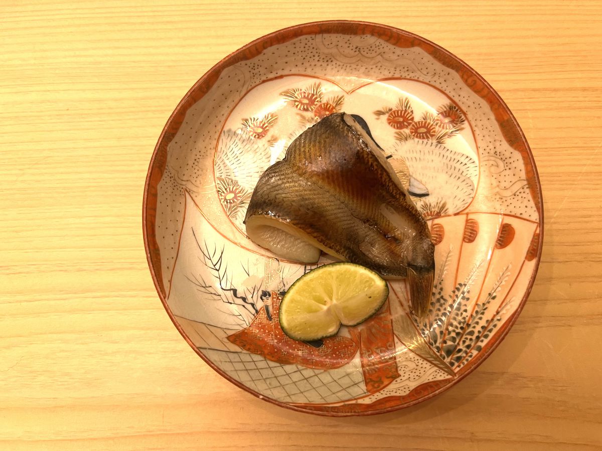 A fish filet sits curled upon itself on a decorative plate; a lime wedge sits on the side.
