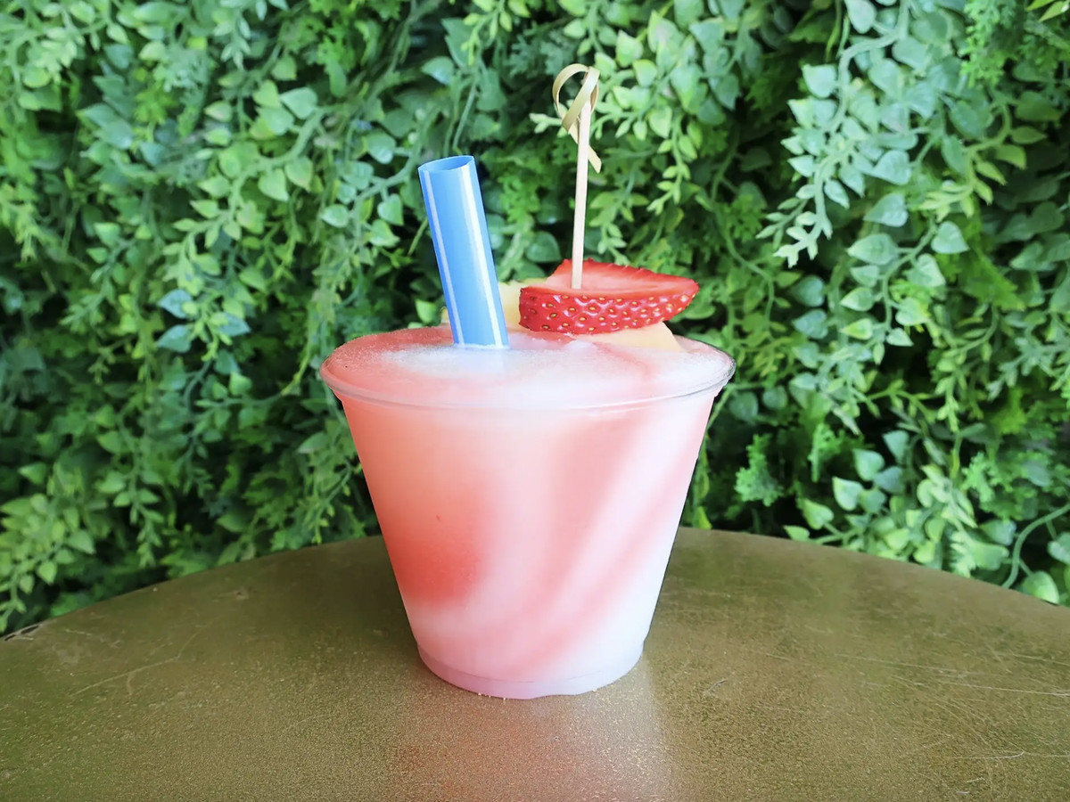 A red and white frozen cocktail sits in a plastic cup on a table in front of a wall of ferns.