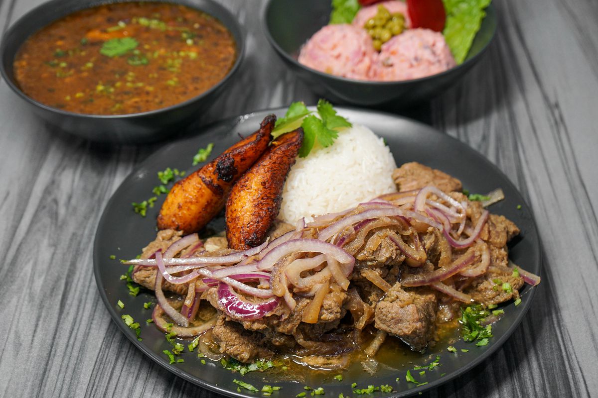Stewed beef covered with red onions and a side of rice with plantains, with pea soup and potato salad to the back.