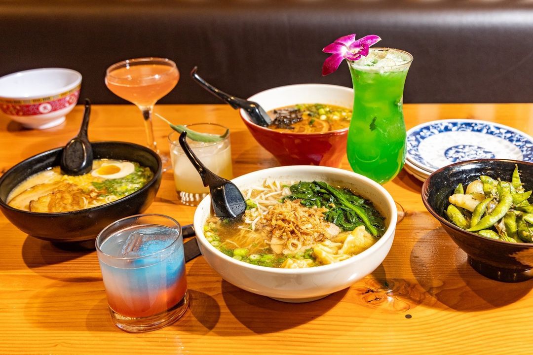 Bowls of ramen, pho, and edamame are spread across a wooden restaurant table alongside colorful cocktails.