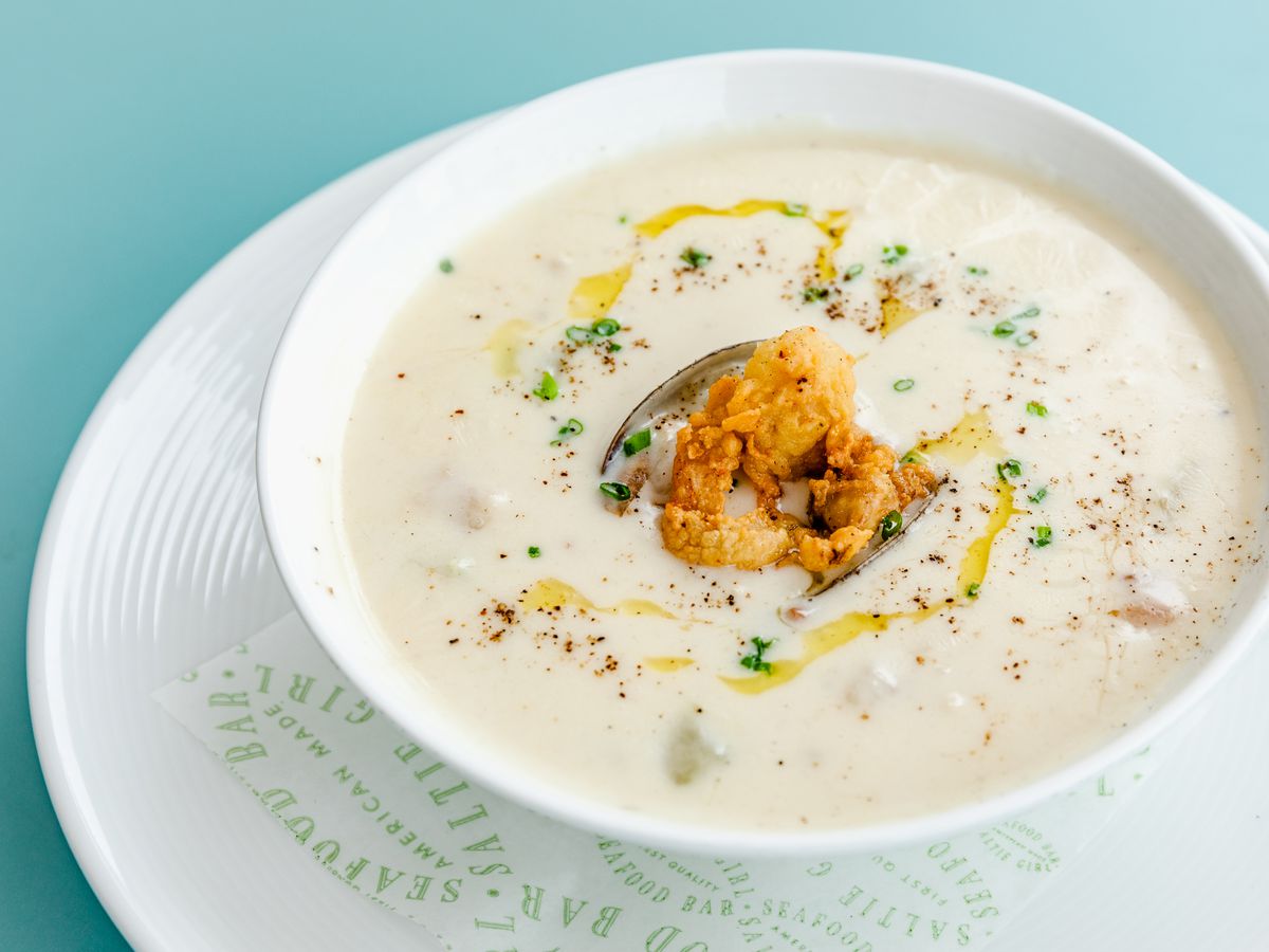 Clam chowder plated in a white cup on a white saucer, garnished with green herbs and fried clams.