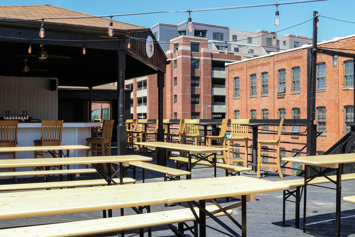 Several long wooden picnic tables accompanied by benches atop a rooftop that has a bar with chairs in the background. Two midrise buildings are in view.