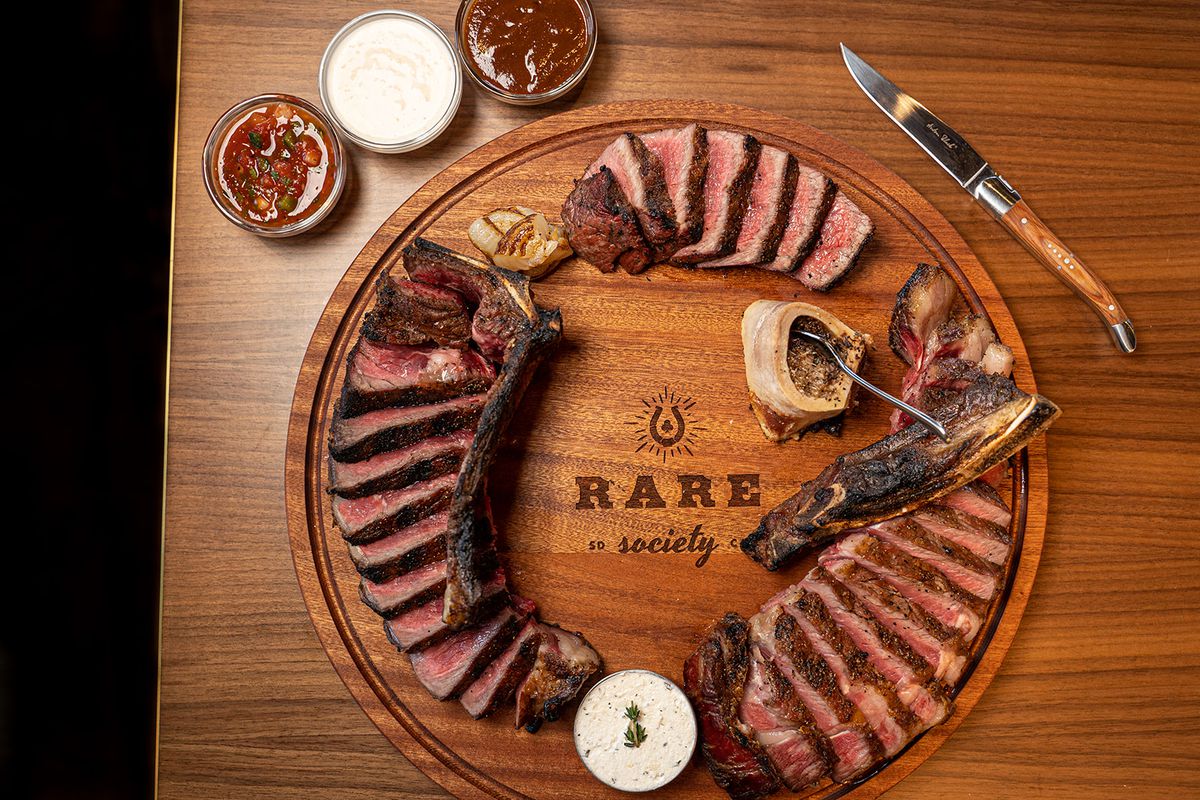 A round wooden board printed with the Rare Society logo with three sliced steaks and small containers of sauce on the side.  