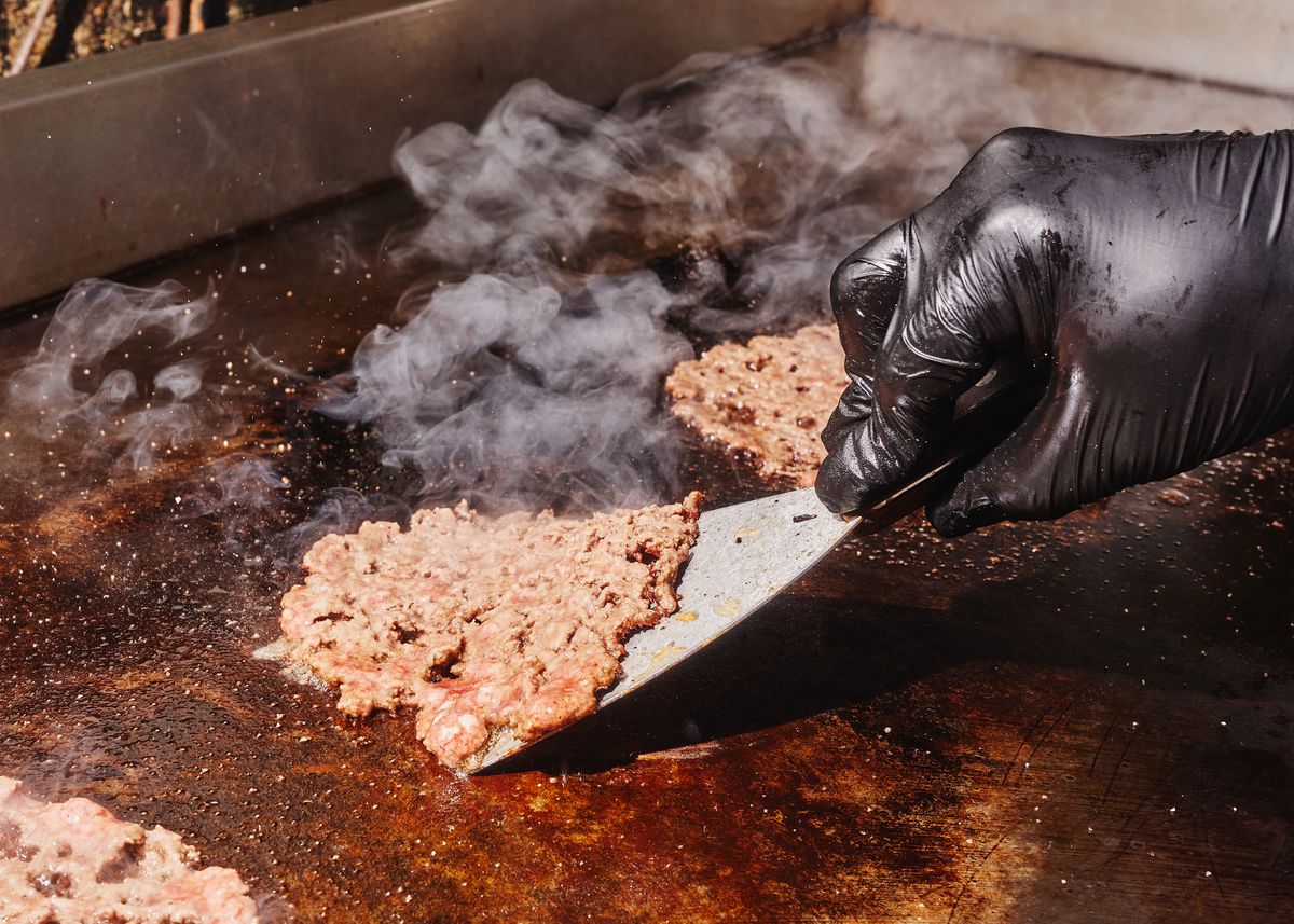 A hand wearing a black glove uses the edge of a spatula to lift up a paper-thin smash burger.