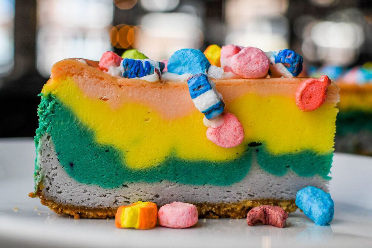 An assault of color in cheesecake form, a tall slice has a graham cracker crust, weird grayish purple marshmallow layer, then stripes of leprechaun green, yellow, and orange. All garnished by tumbles of Lucky Charms marshmallows