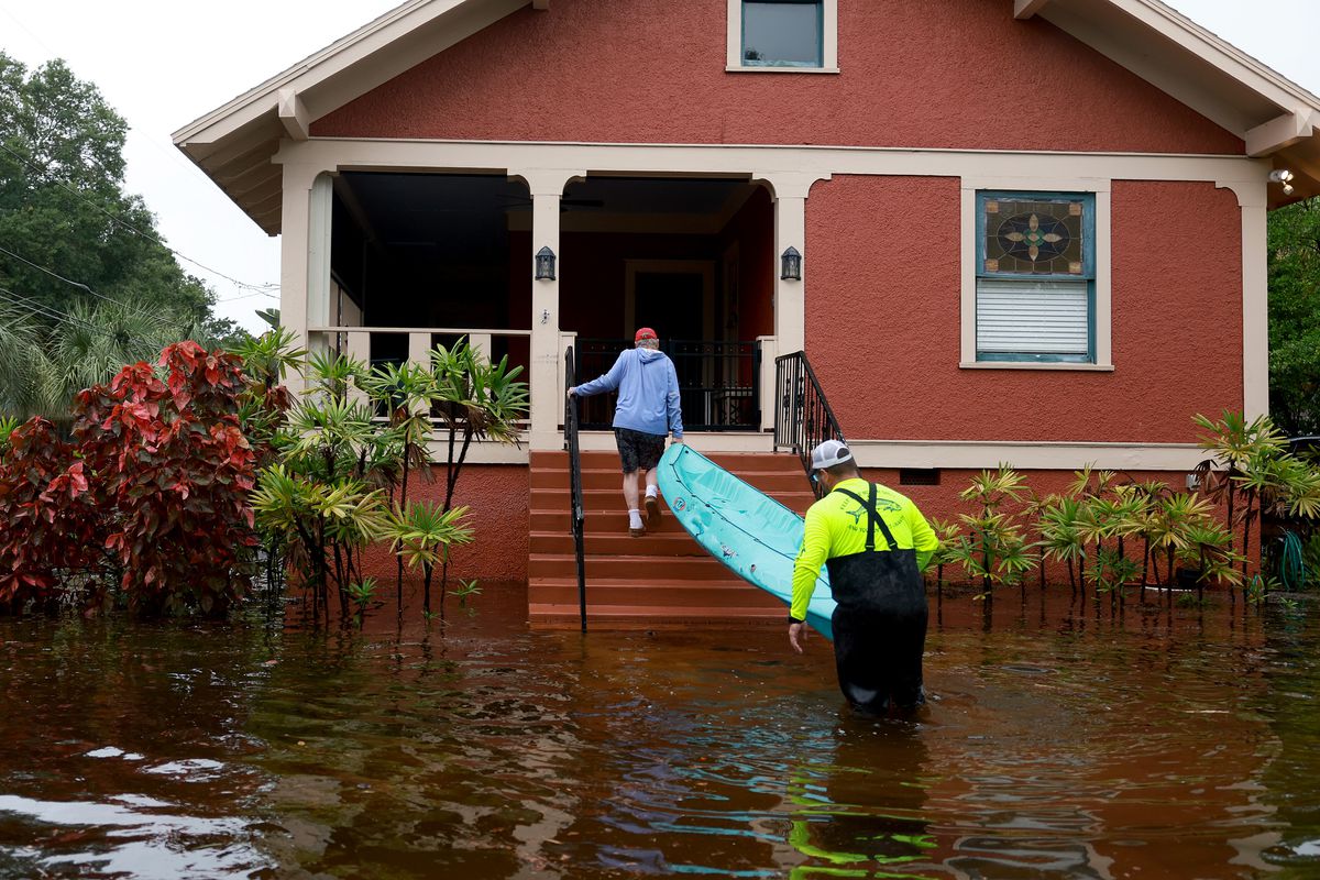 One person walks up the porch stairs to a house while holding one end of a kayak, and another person holds the other end of the raft, still standing in the flooded water where a front lawn would be.