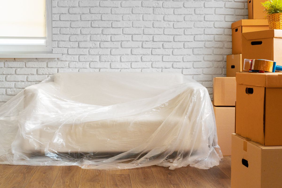 A white couch inside a home with clear wrapping over it, brown cardboard moving boxes, and packing tape. 
