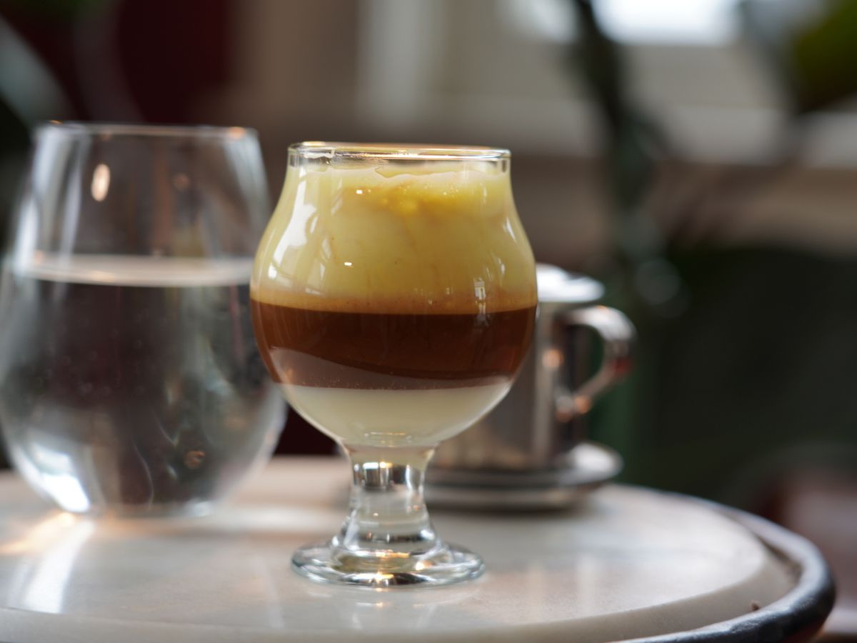 A three-layered coffee drink sits in a curved glass on a white table. There’s a milk layer, a coffee layer, and a foam layer visible.