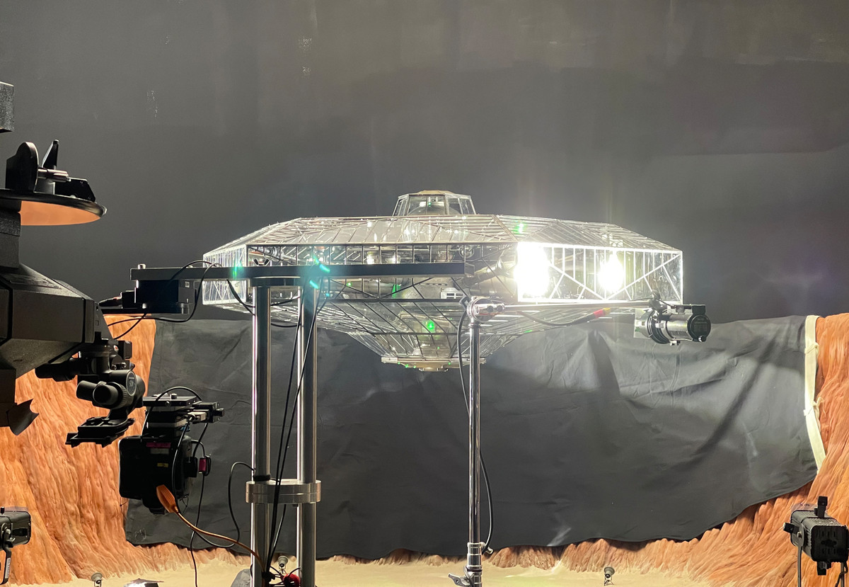 A transparent, boxy model spaceship is held up on a metal frame, with lights and cameras around it.