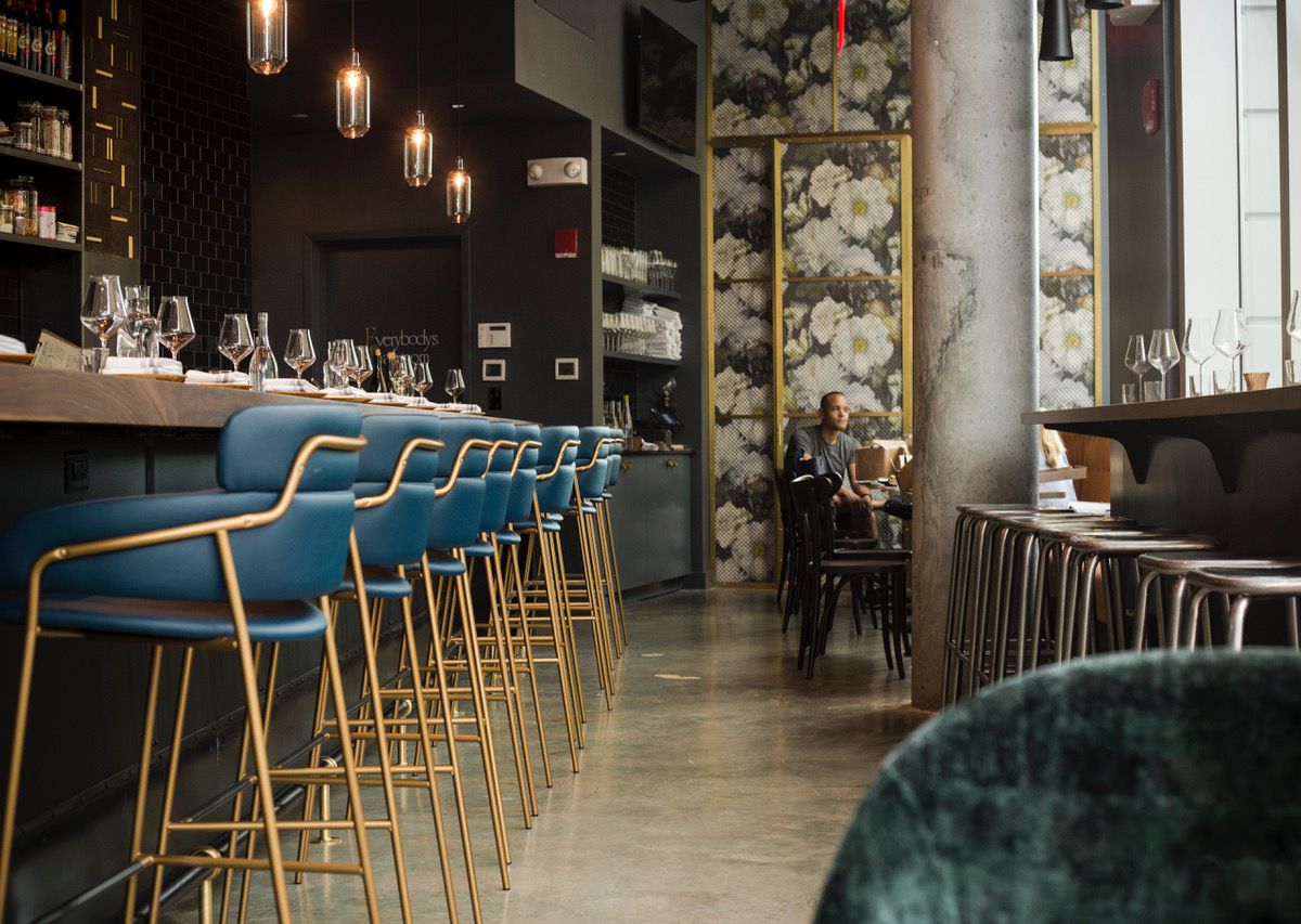 Blue and gold bar stools line a dark brown bar, with backless silver stools arranged at a window counter.