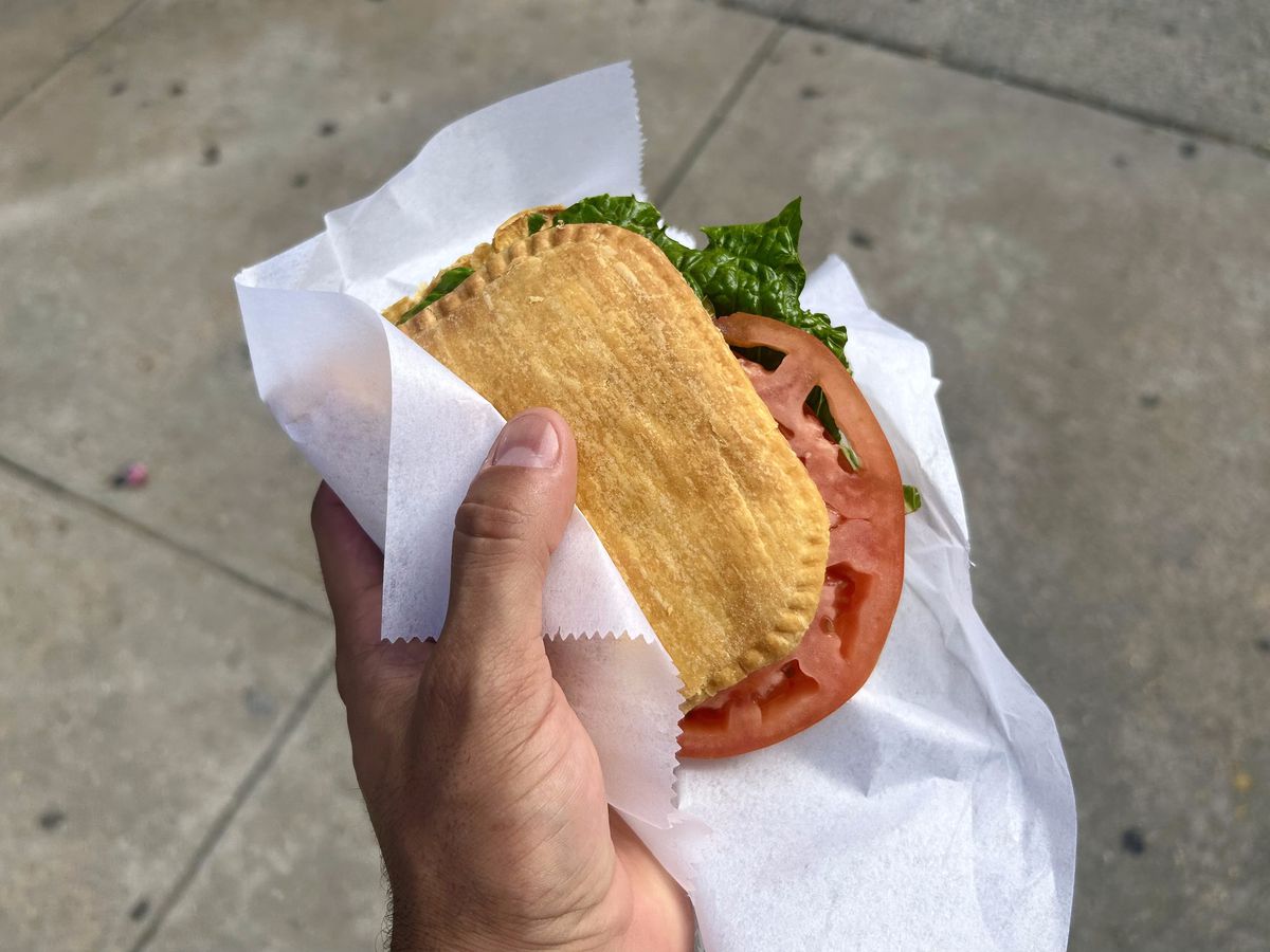 A hand holds a Jamaican beef patty with lettuce and tomato.