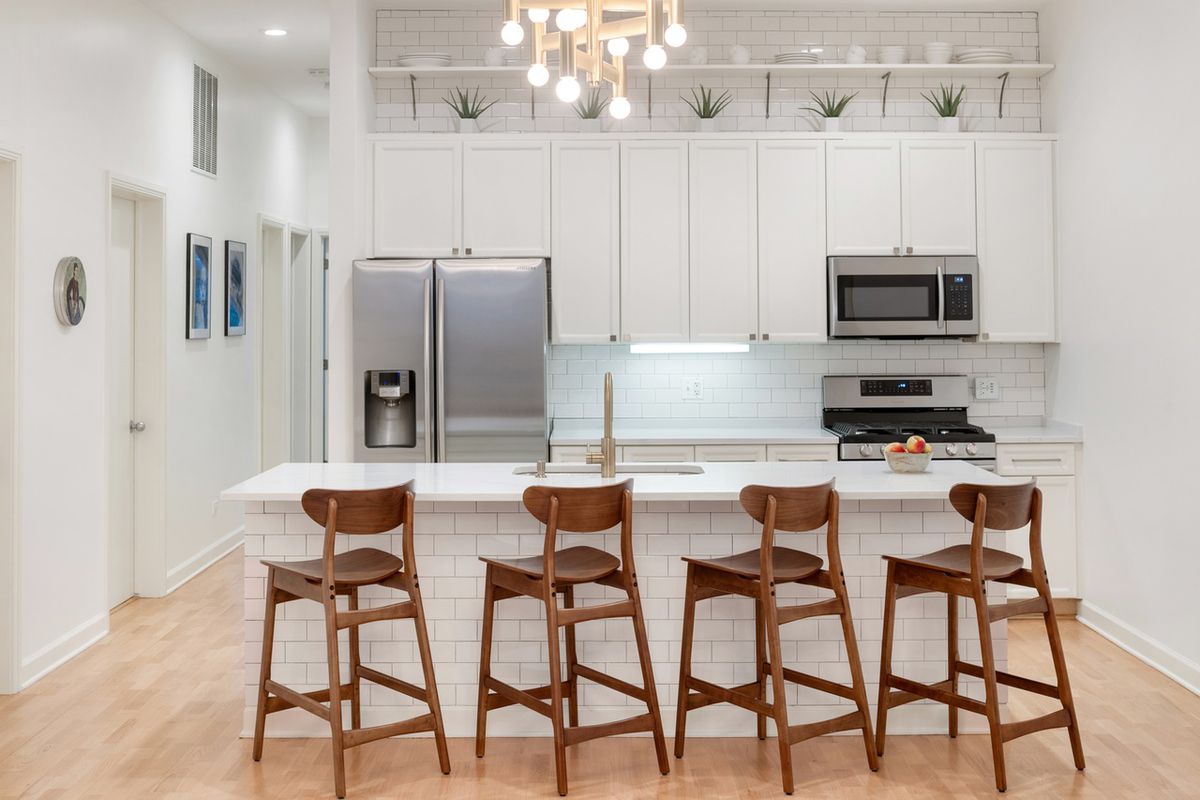 A compact white-colored kitchen with white subways tiles on the backsplash and central island, that has four wooden stools. 