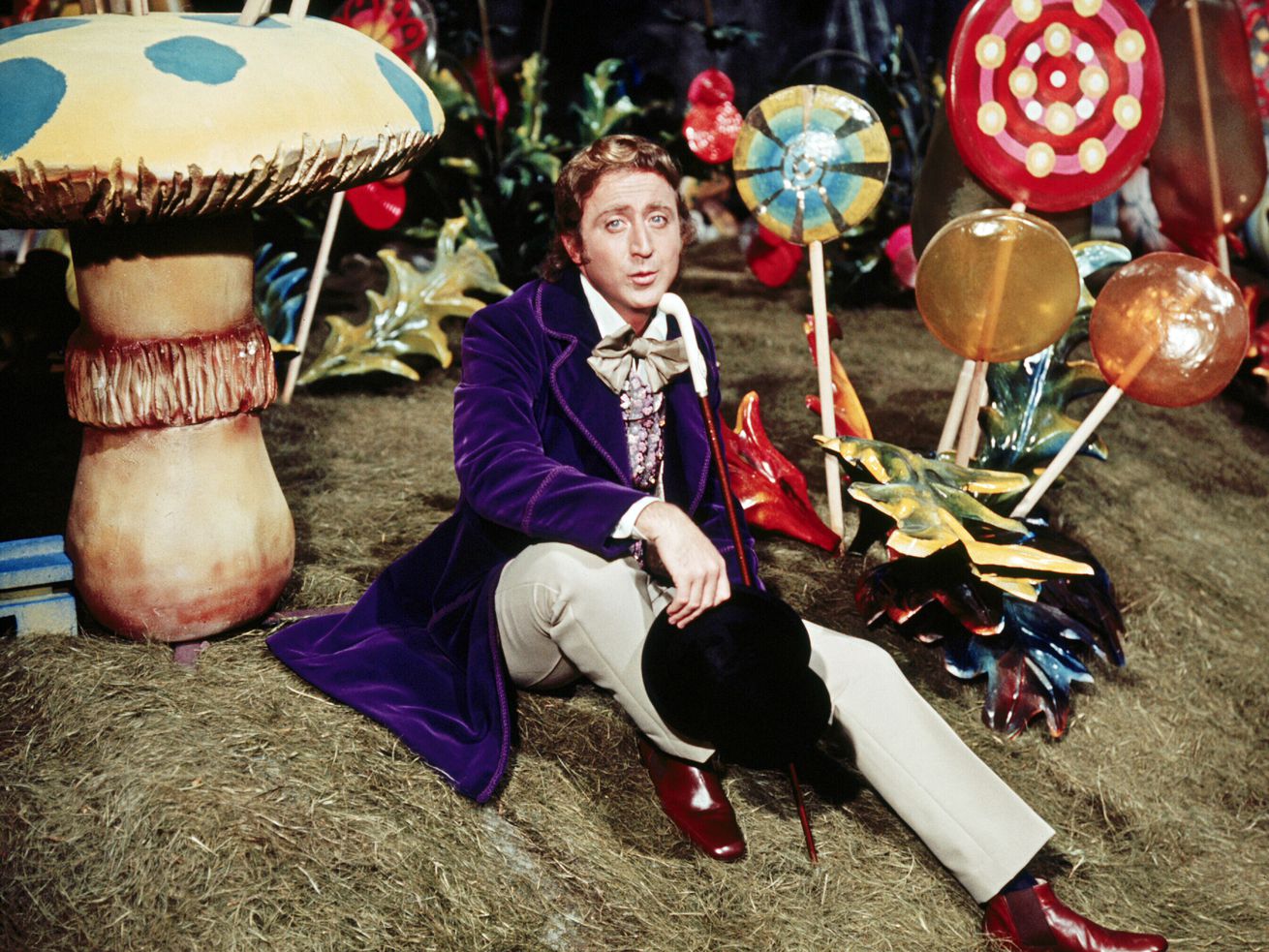 Wilder dressed in a purple coat as Wonka sits among oversize lollipops and mushrooms.