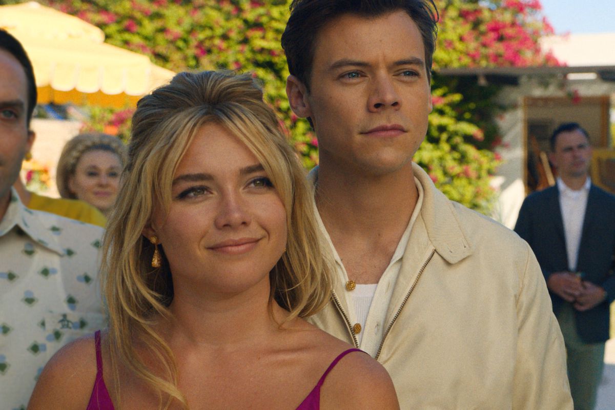 Florence Pugh as Alice and Harry Styles as Jack smile at a sunny garden party, surrounded by other characters, in Don’t Worry Darling.