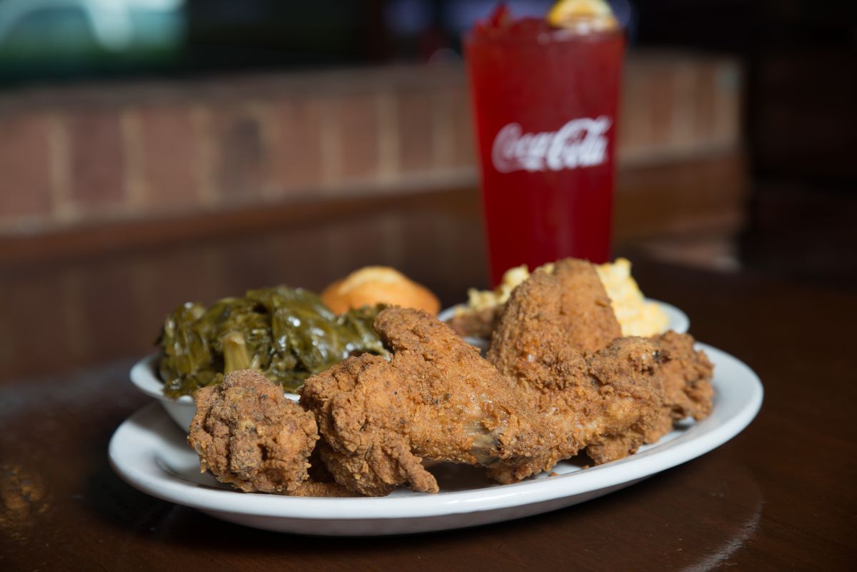 <p style="user-select: auto;">A white dish with fried chicken pieces, collared greens, and a corn muffin with a glass full of soda.