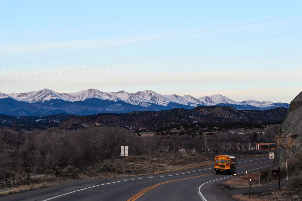 A yellow school bus drives around a curve in the highway with a view of snow-capped mountains and blue skies in the background.