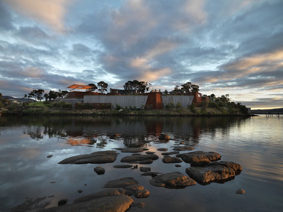 The buildings of the MONA museum on an islet at sunset with water all around, small stones poking out of the water, and pillowy clouds against a blue sky.