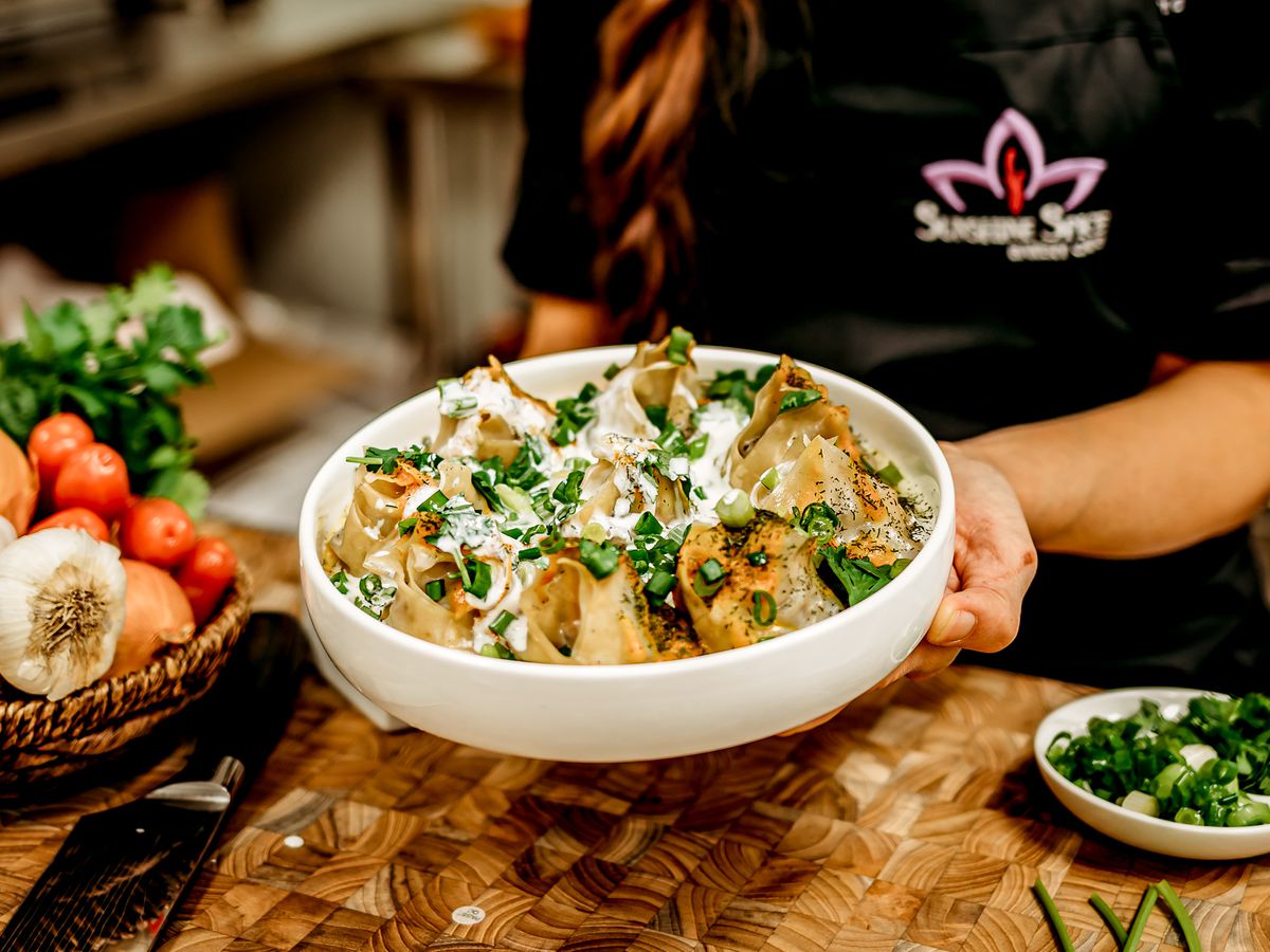 A server holds a bowl of dumplings topped with cream, shopped scallions, and herbs.