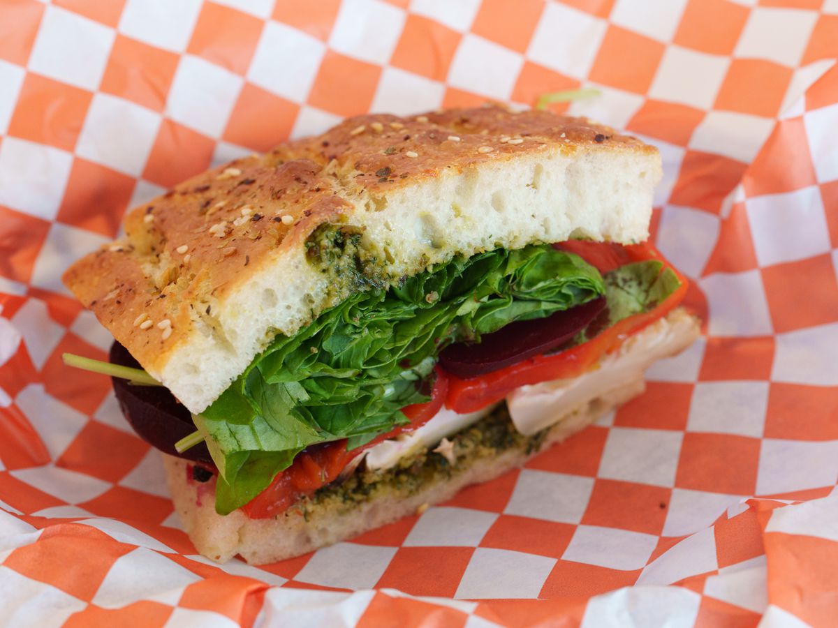 A focaccia sandwich with lettuce, tomatoes, tofu, and pesto on red and white checkered paper.
