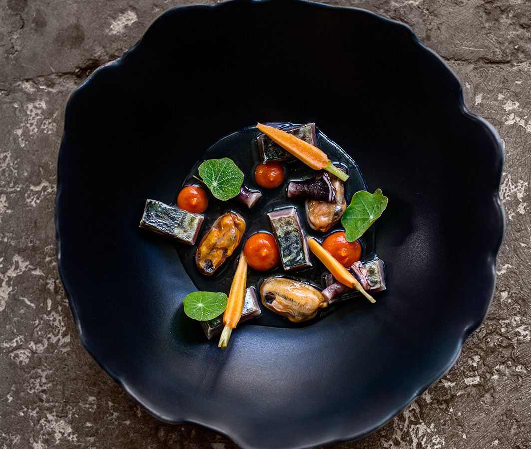 A dark bowl with textured edges. Inside a mix mussels, vegetables, and herbs