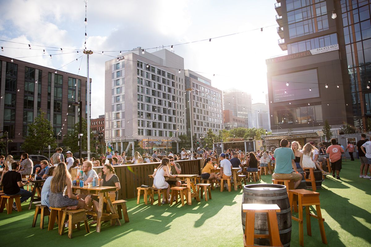 Crowds of people in summer clothing sit at picnic tables on astro turf in a large city beer garden on a sunny day.