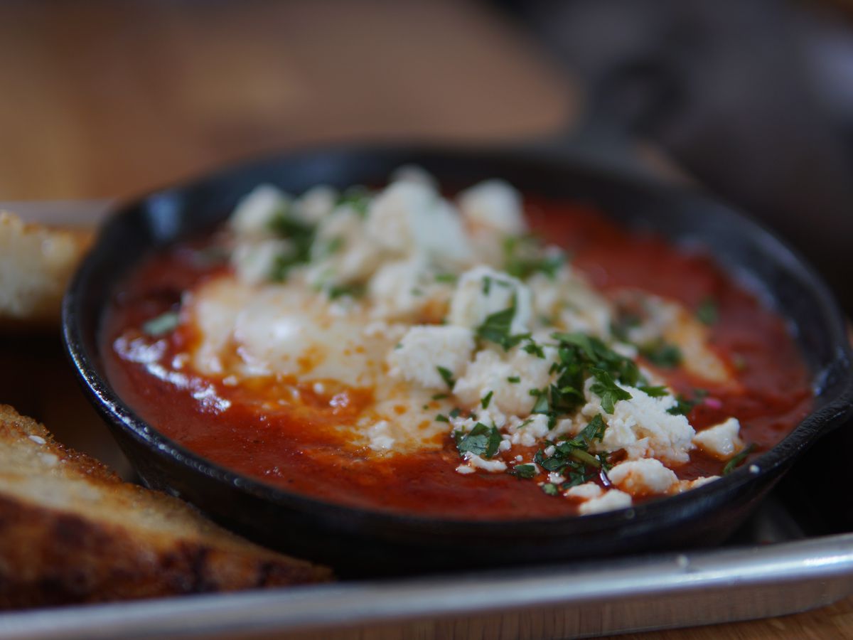 A fire engine red shakshuka sits in a cast iron with crumbles of feta and herbs over poached eggs