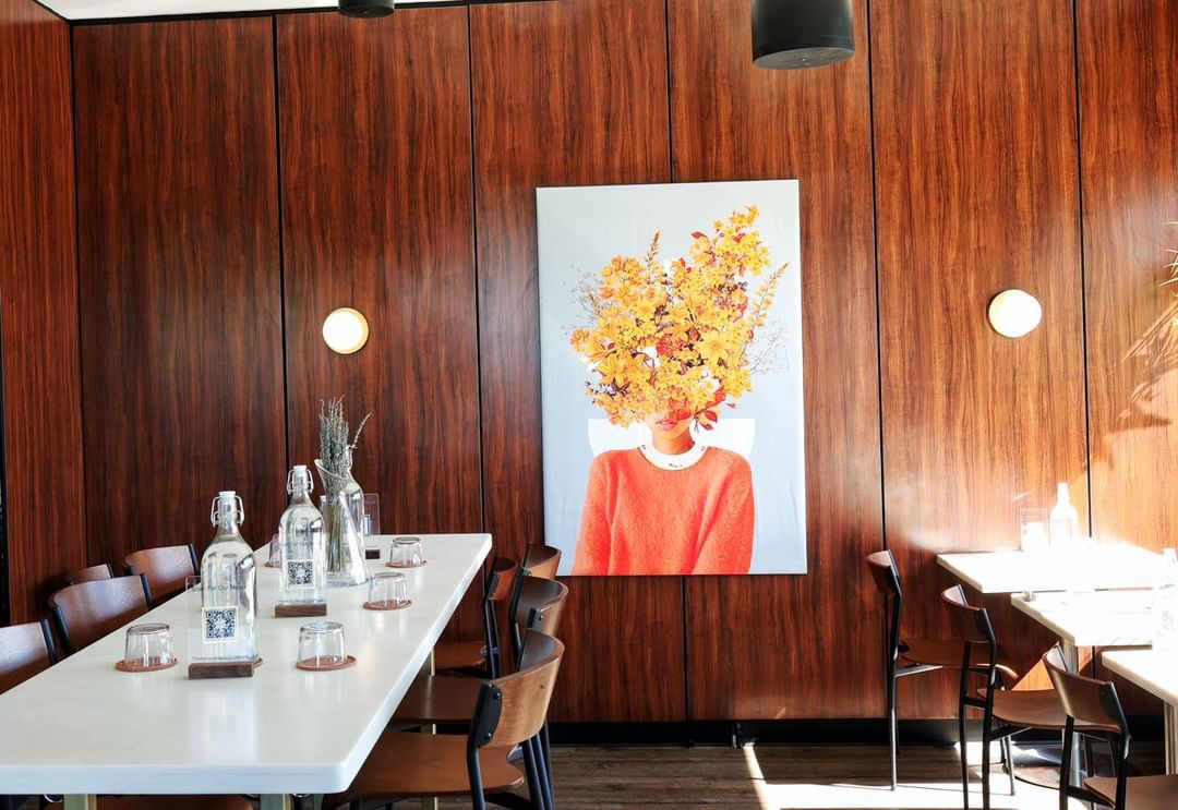 A wood-lined dining room with long white tables set with water pitchers, and a large piece of art on the wall depicting a person with a large burst of flowers for a head. 