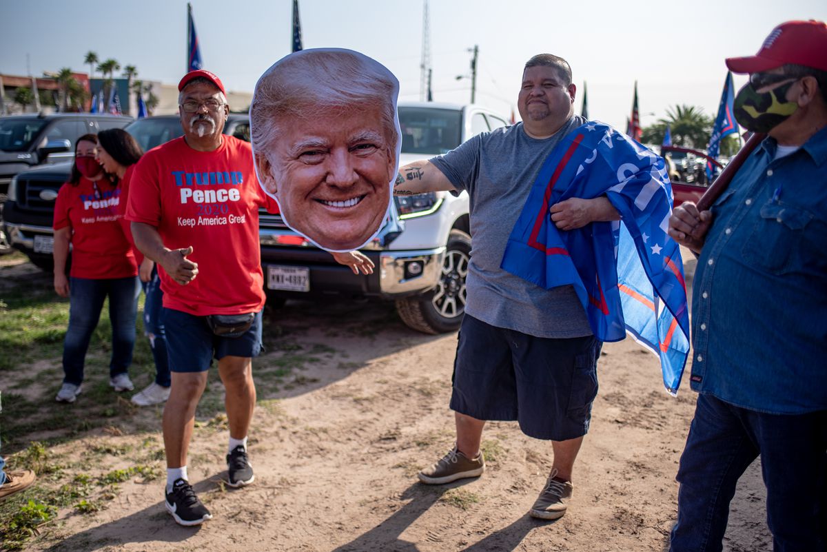 A Latino man draped in a blue Trump-Pence flag holds a giant cut out of President Donald Trump’s head. Behind him, a white haired Latino man in a red Trump-Pence shirt gives a thumbs up. They stand in a sunny field full of trucks bearing Trump flags.