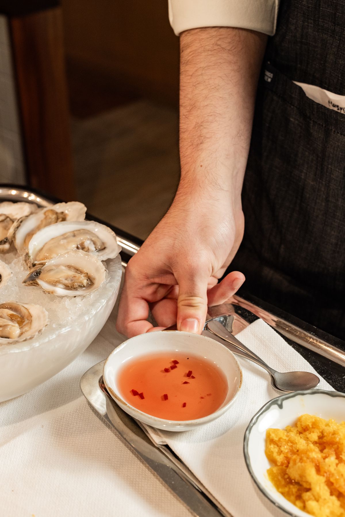 A man’s hand holds a bowl of chili sauce. To the right is a frozen ganache. To the left is a bowl of iced, opened oysters.