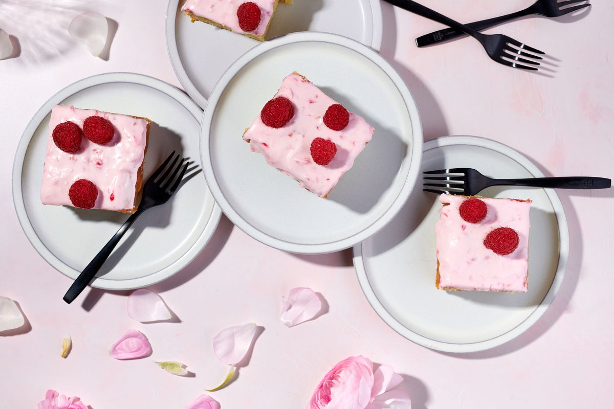 Square slices of raspberry cardamom cake arranged on small plates, against a pink backdrop scattered with pink petals.