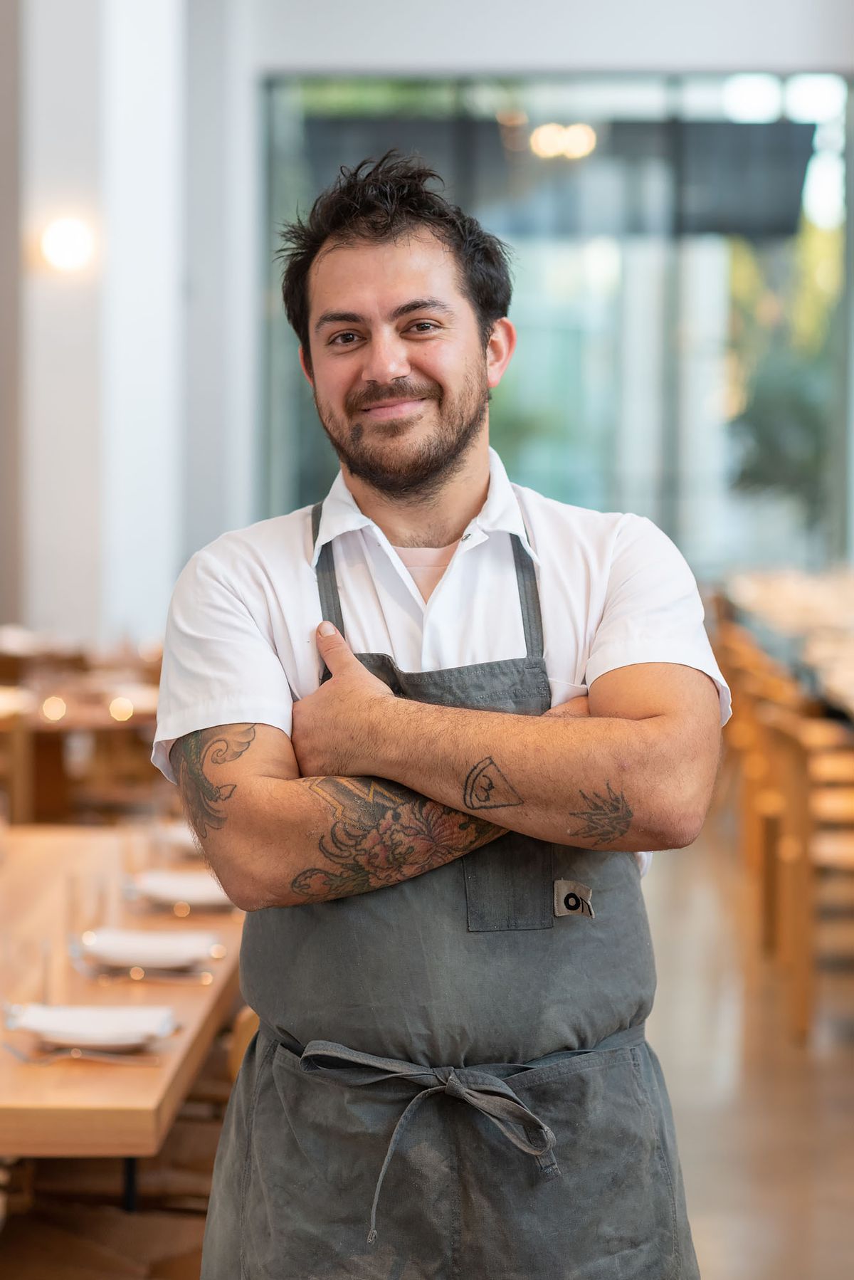 A man in a chef’s apron stands with his arms crossed, smiling.