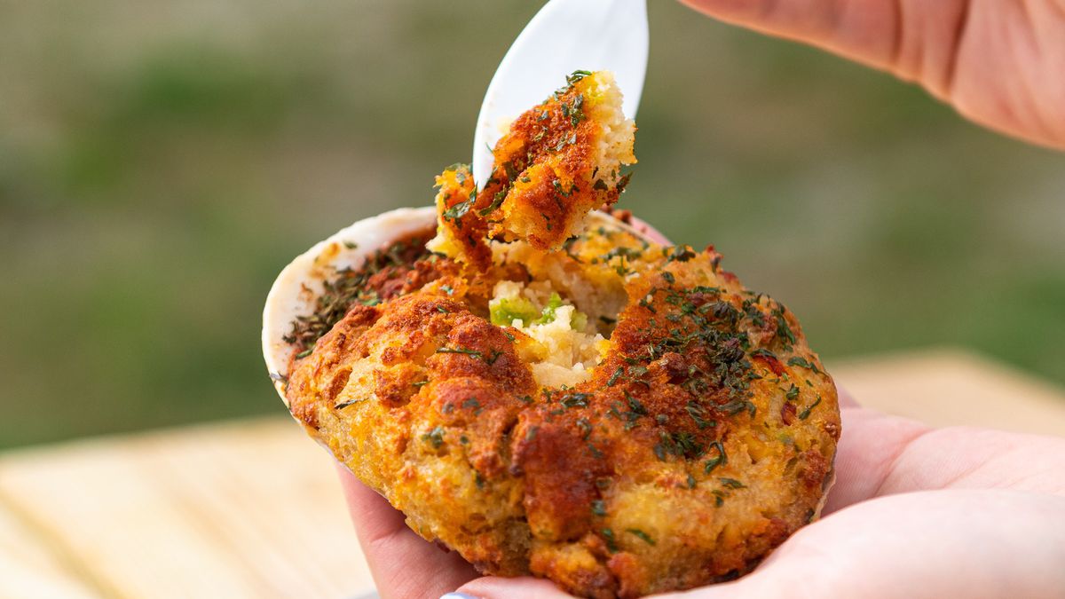 A hand holds up a stuffed quahog shell, using a plastic spoon to grab some of the stuffing.