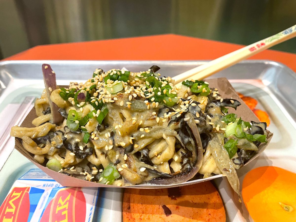 A paper container filled with fried noodlese with vegetables, mushrooms, and cabbage with a pair of chopsticks.