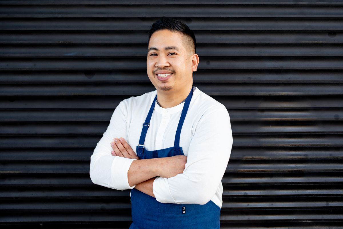 A man in a white shirt with a blue apron smiles and crosses his arms. 