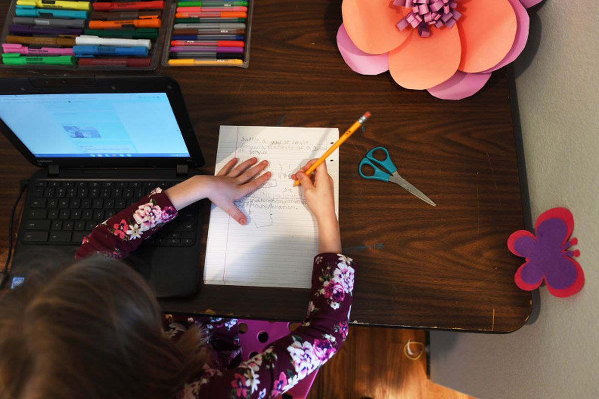 A girl wearing a colorful shirt works from her desk at home, writing on a piece of paper next to her laptop.