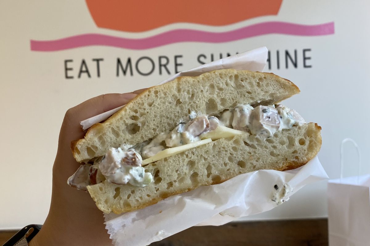 A hand holds up a half of a sandwich with fillings exposed against a wall with “Shirley Eat More Sunshine” in the background.