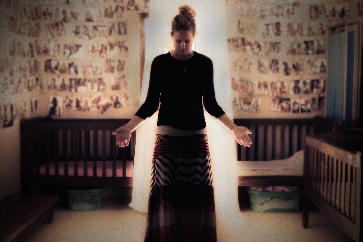 A young woman stands in the middle of a room, head bowed, arms outstretched.